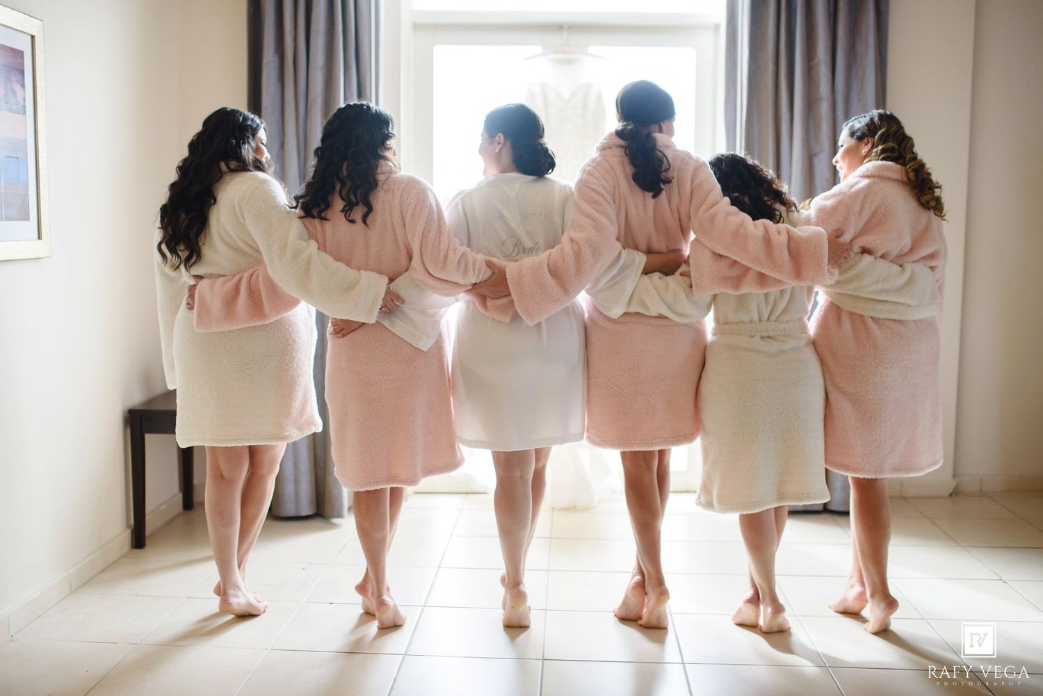 Bridesmaids in matching pink and white robes standing together with arms around each other, viewed from behind.