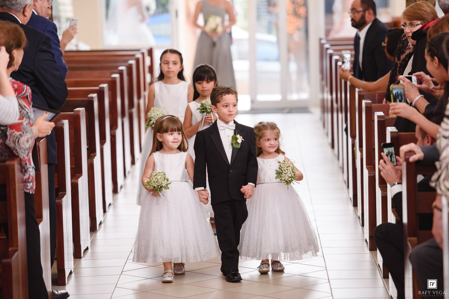 Flower girls and ring bearer walk down church aisle during wedding ceremony.