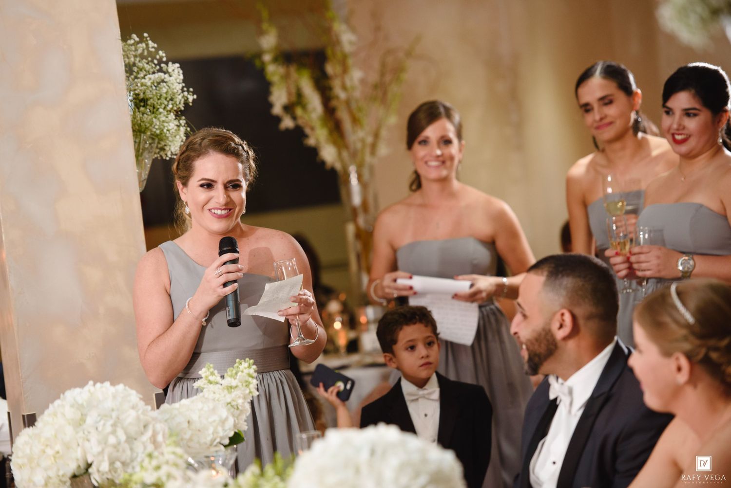 Bridesmaid delivering a heartfelt speech at an elegant wedding reception with floral arrangements and bridal party in gray dresses.