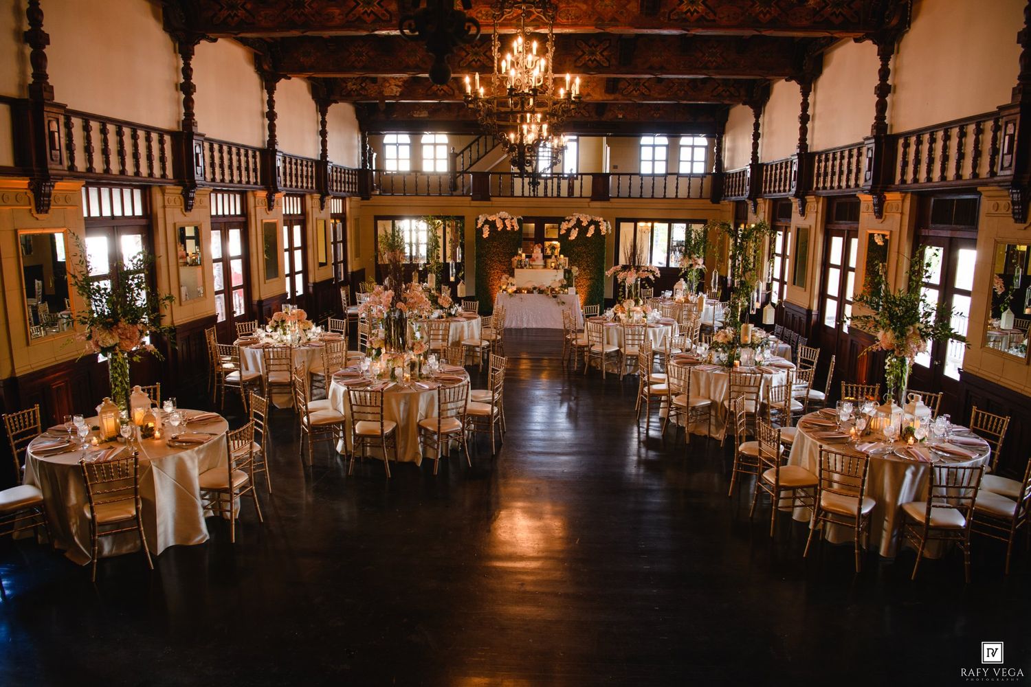 Grand wedding reception hall with dark wood floors, chandeliers, gold chiavari chairs, and beautifully decorated round tables.