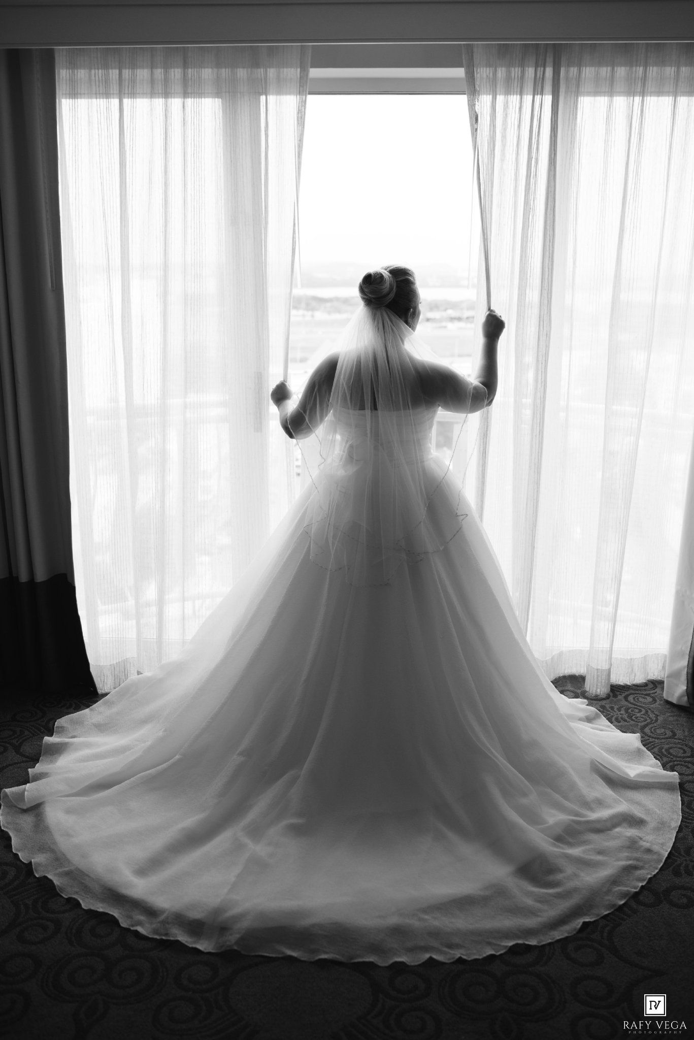 Bride in a ballgown wedding dress with veil stands at a window, viewed from behind in black and white.