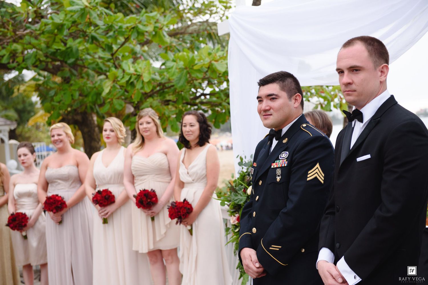 Military groom and groomsmen stand at altar with bridesmaids in champagne dresses holding red roses at outdoor wedding.