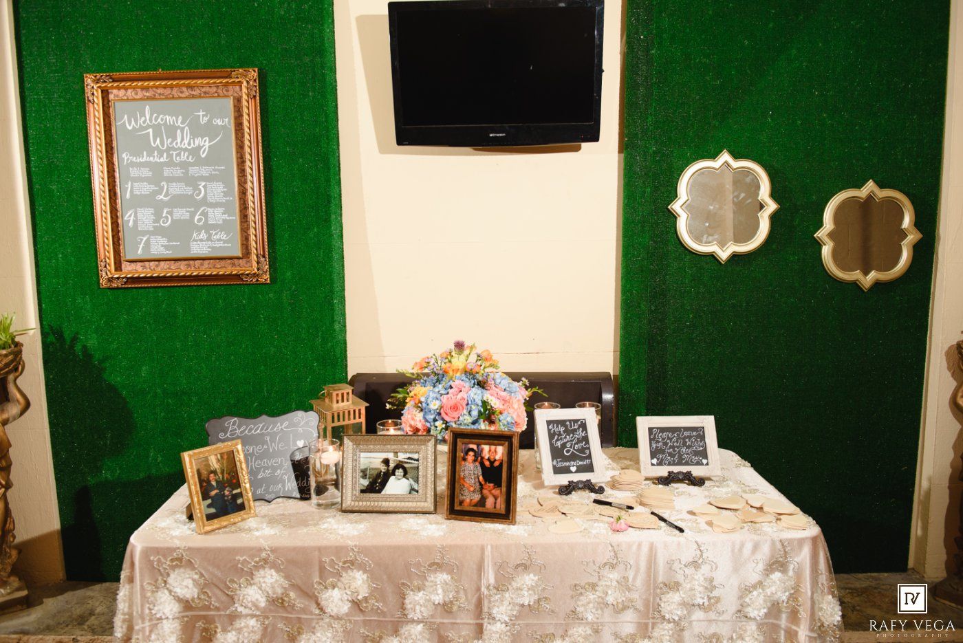 Wedding memory table displaying framed photos, flowers, and guest book against a green moss wall backdrop.