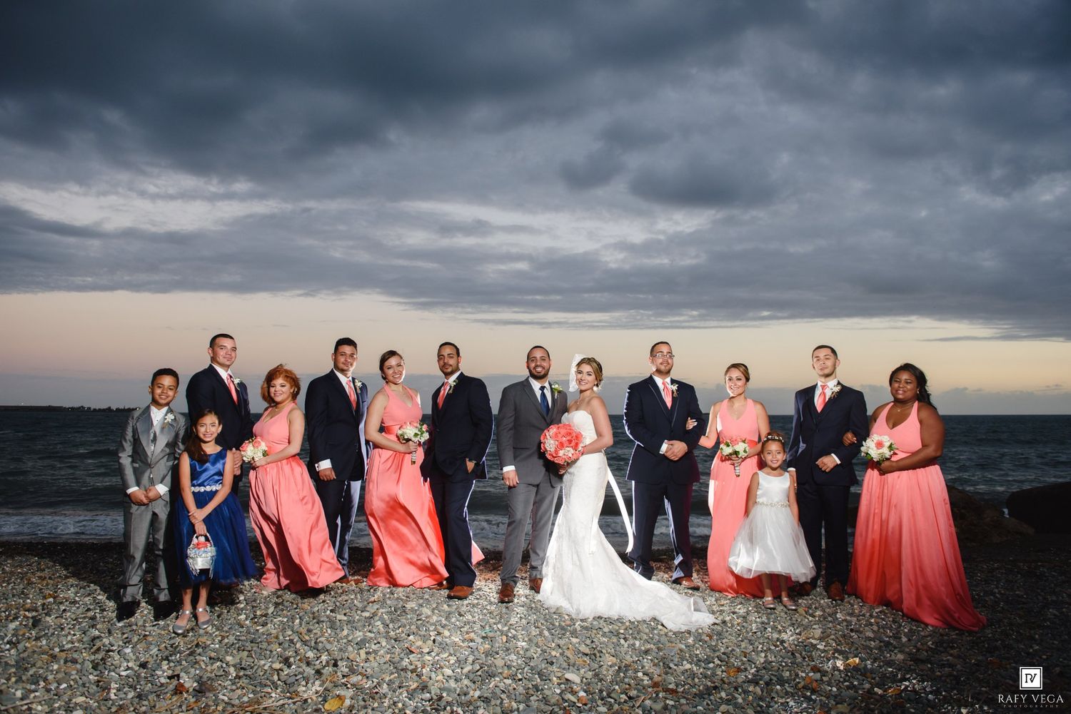 Wedding party poses on rocky beach shore with dramatic cloudy sky, bride in white gown with coral-dressed bridesmaids.