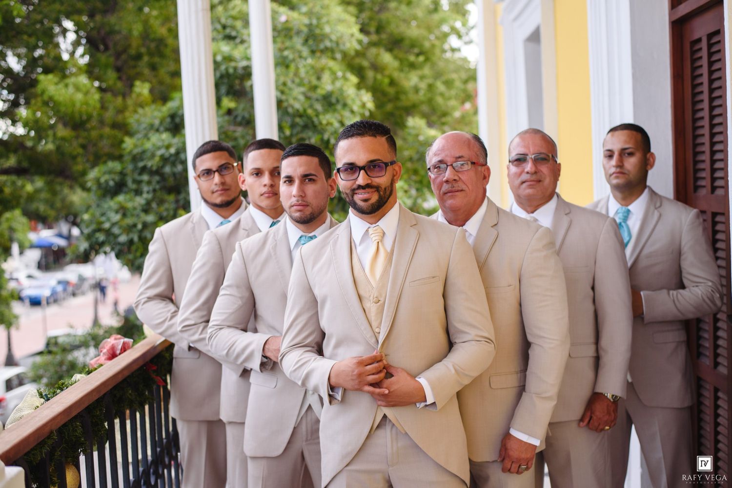 Groomsmen in tan suits pose on a balcony with the groom wearing sunglasses and a yellow tie at a wedding.
