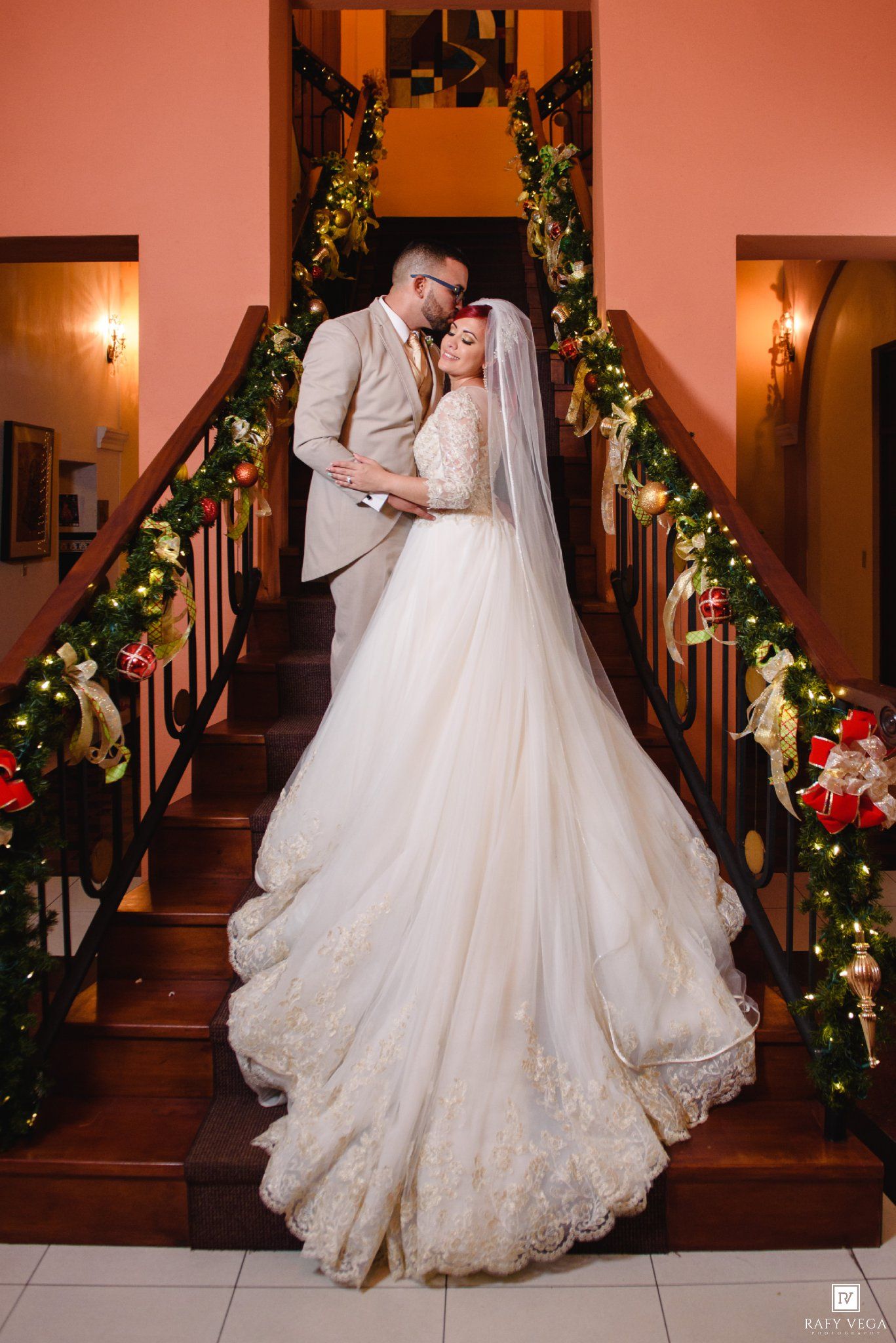 Bride and groom share romantic moment on Christmas-decorated staircase, bride in elegant lace ballgown with veil.