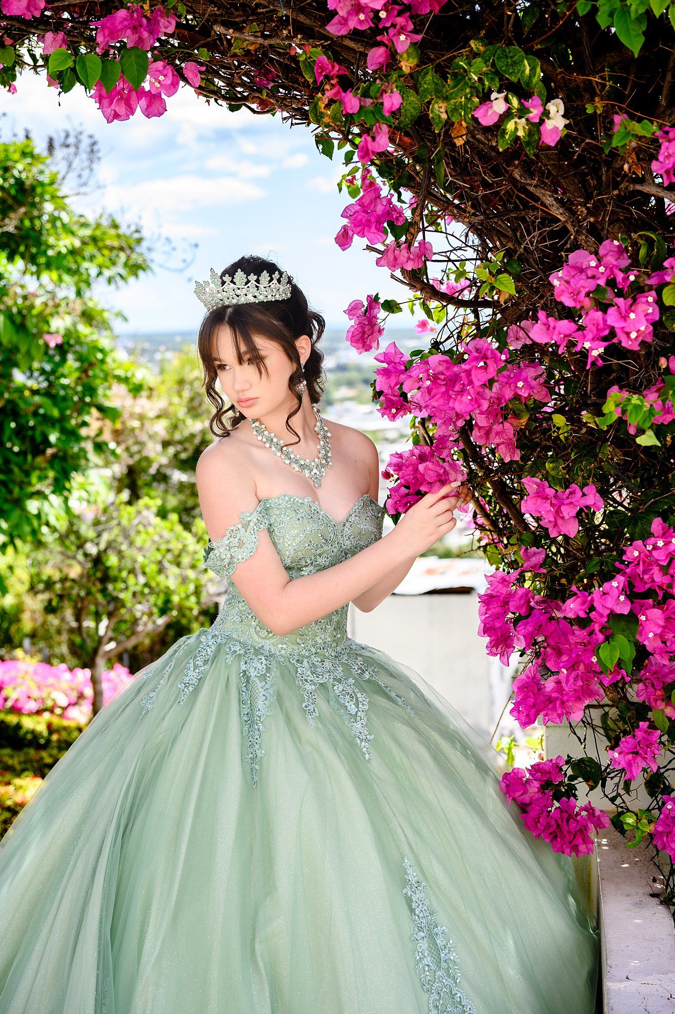 Young woman in sage green ball gown and tiara poses beside vibrant pink bougainvillea flowers in a garden.