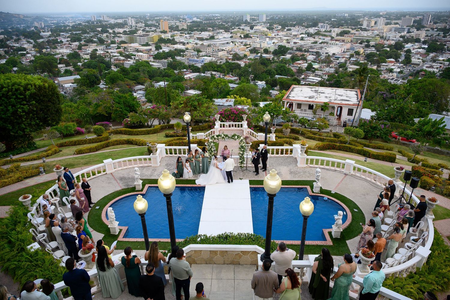 Elegant outdoor wedding ceremony on a terrace overlooking a city, with guests seated around a pool and floral arch altar.