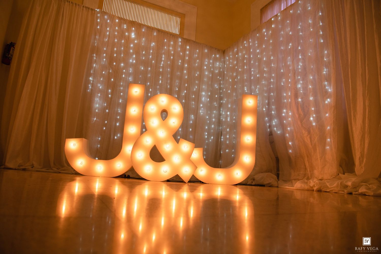 Illuminated marquee letters J&J with warm bulb lights against a sparkling fairy light backdrop at a wedding reception.