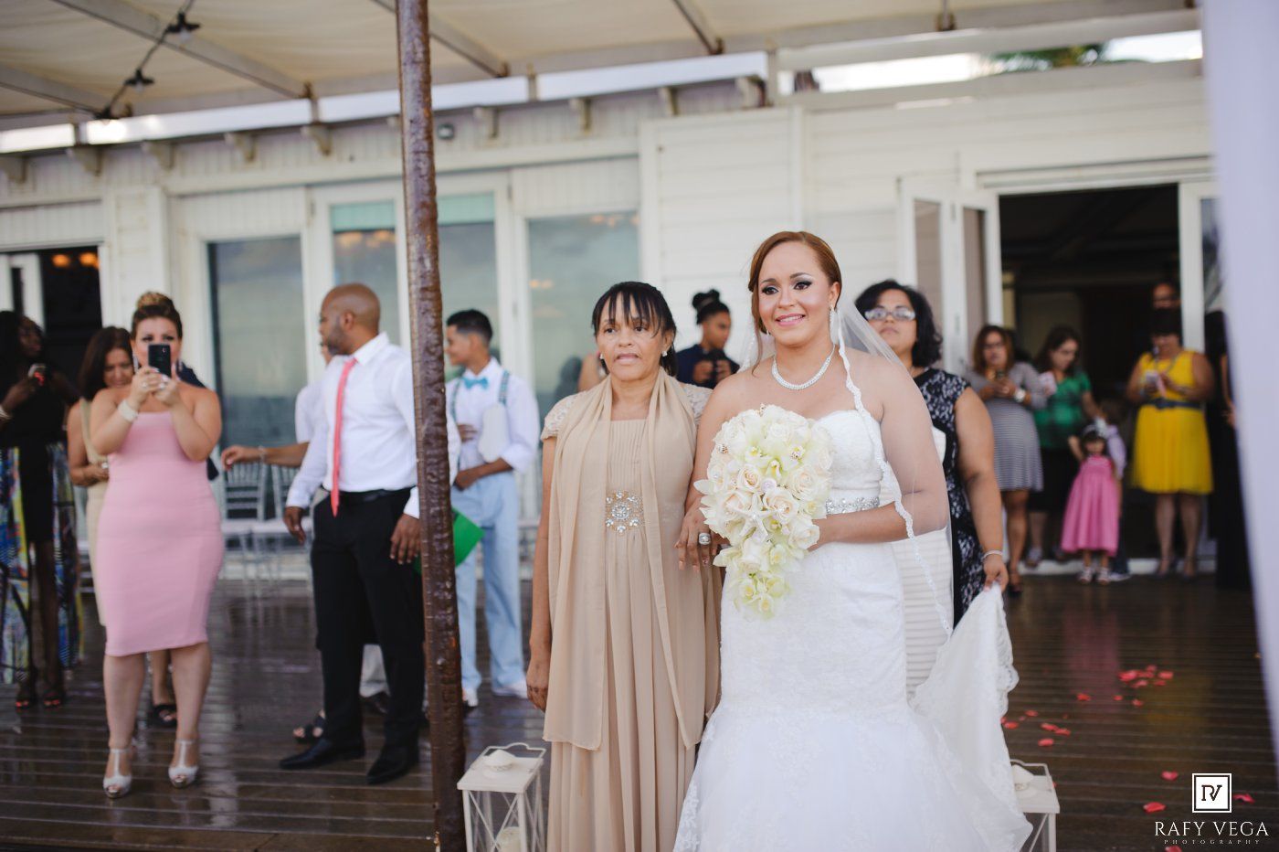 Bride in white gown holding bouquet walks down aisle with escort at outdoor wedding ceremony.