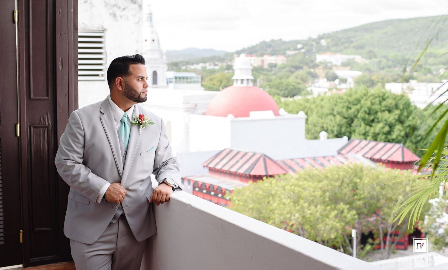 Groom in light gray suit with teal tie and boutonniere stands on balcony overlooking scenic cityscape with red dome.