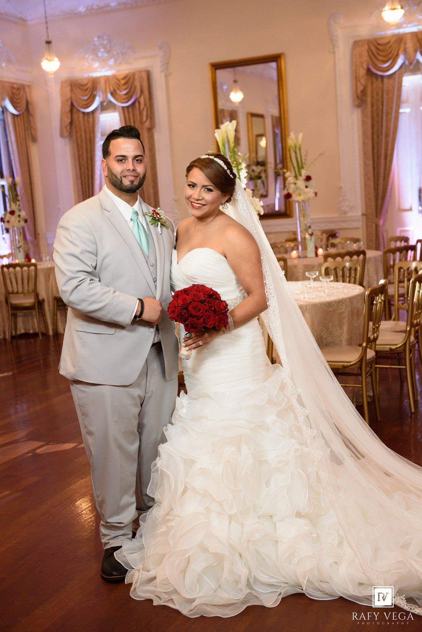 Bride in white ruffled gown with red bouquet and groom in gray suit pose together in elegant wedding venue.