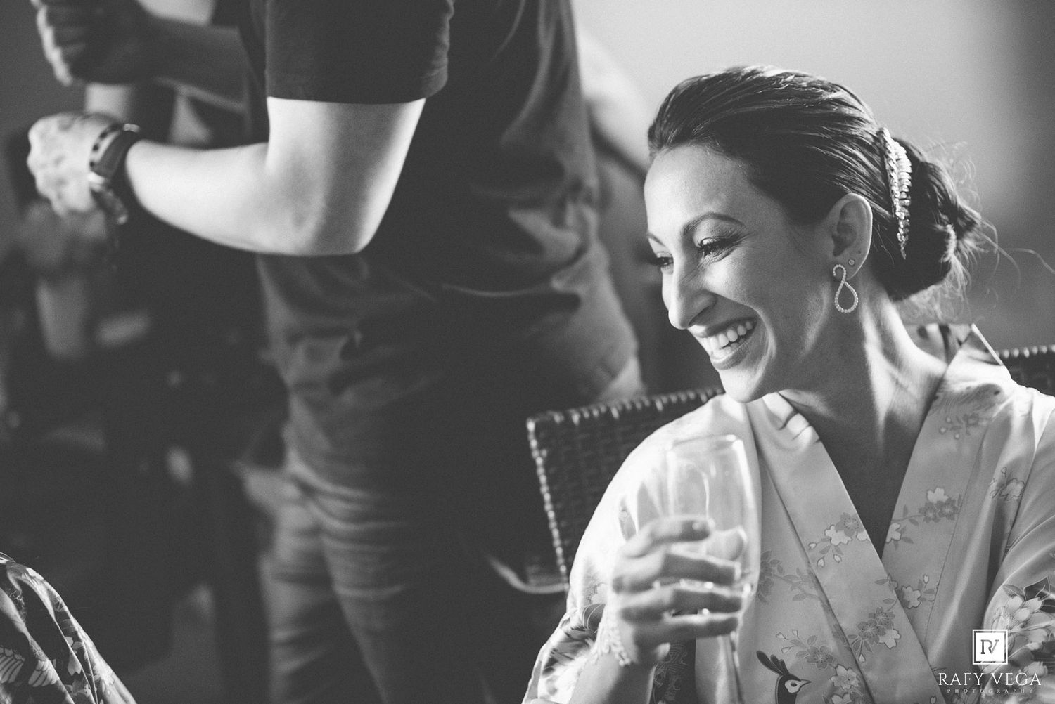 Black and white photo of a smiling bride in a robe holding a glass during wedding preparations.