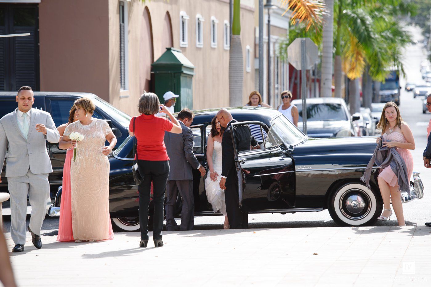 Elegantly dressed group near a classic black car on a sunny street, suggesting a formal event or wedding.