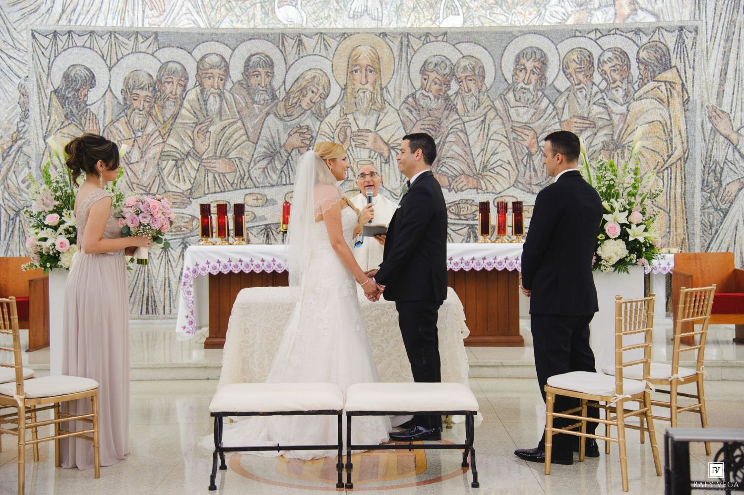 Bride and groom exchange vows at elegant church altar with mosaic backdrop, flanked by officiant and bridesmaid.