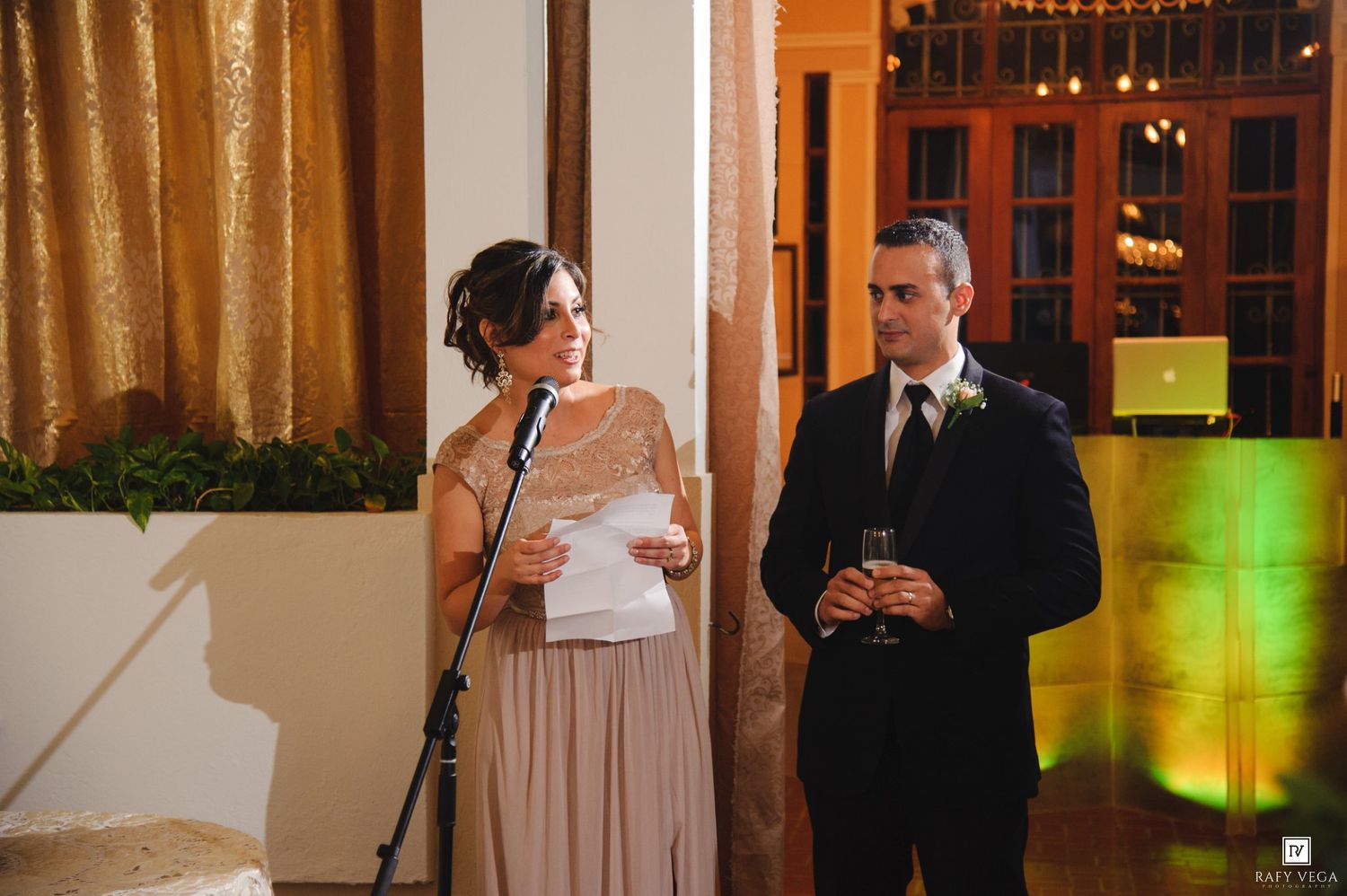 Woman reads speech at microphone beside man in tuxedo at elegant wedding reception with gold curtain backdrop.