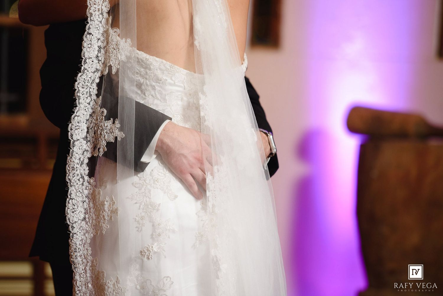 Bride and groom holding hands during wedding ceremony, bride wearing lace gown and veil, purple uplighting in background.