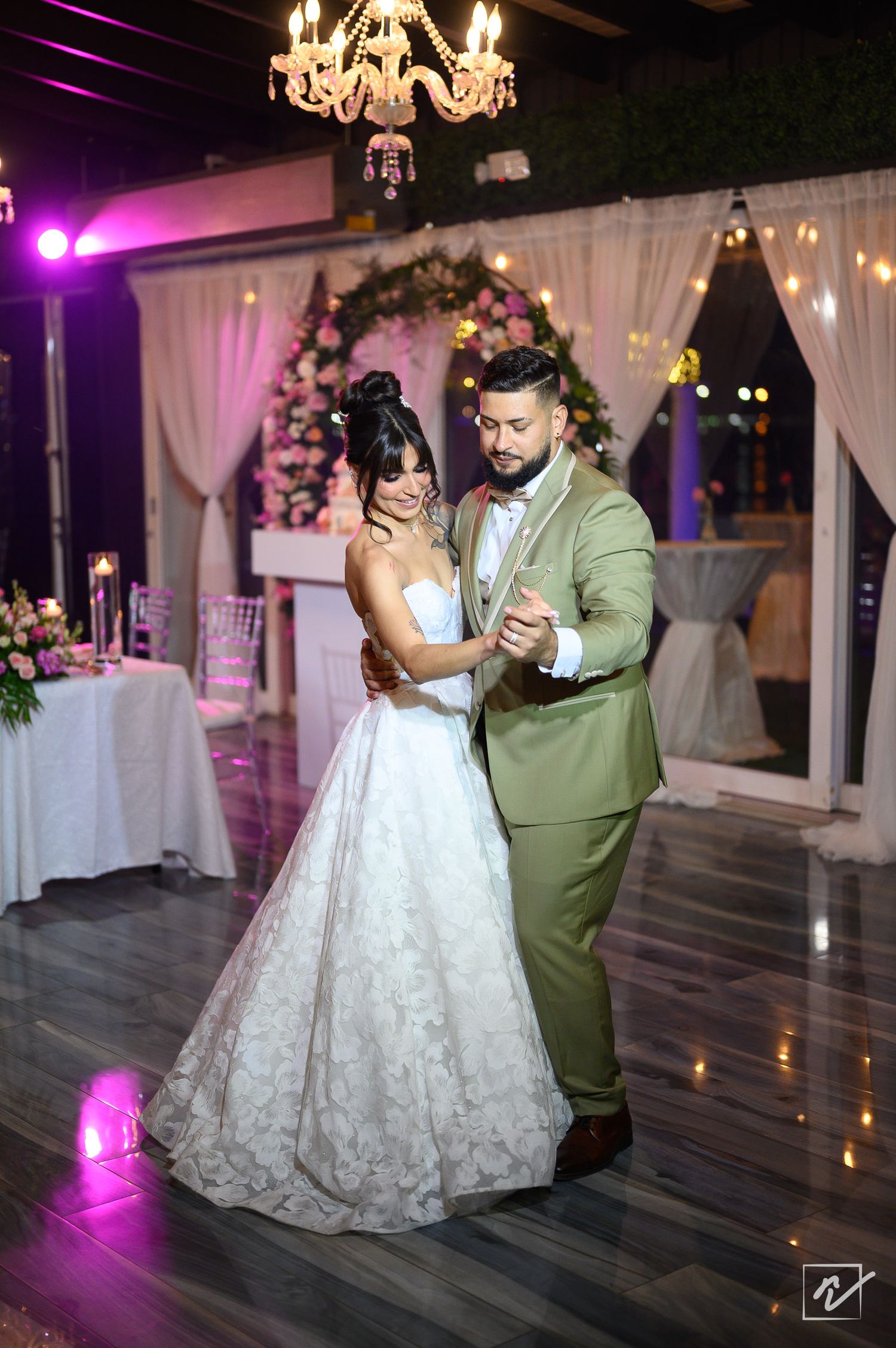Bride in a lace wedding gown and groom in a green suit share a first dance under a floral arch and chandelier at a reception.
