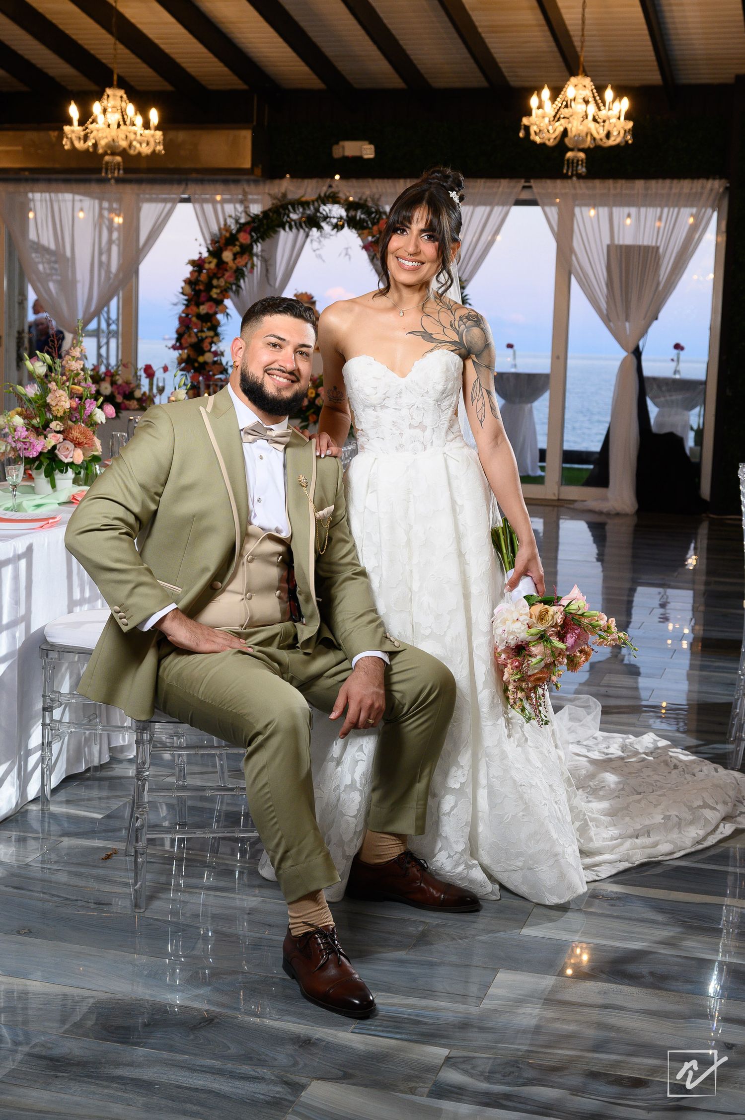 Smiling bride in a lace strapless gown stands beside a seated groom in an olive green suit at an elegant wedding venue with chandeliers.