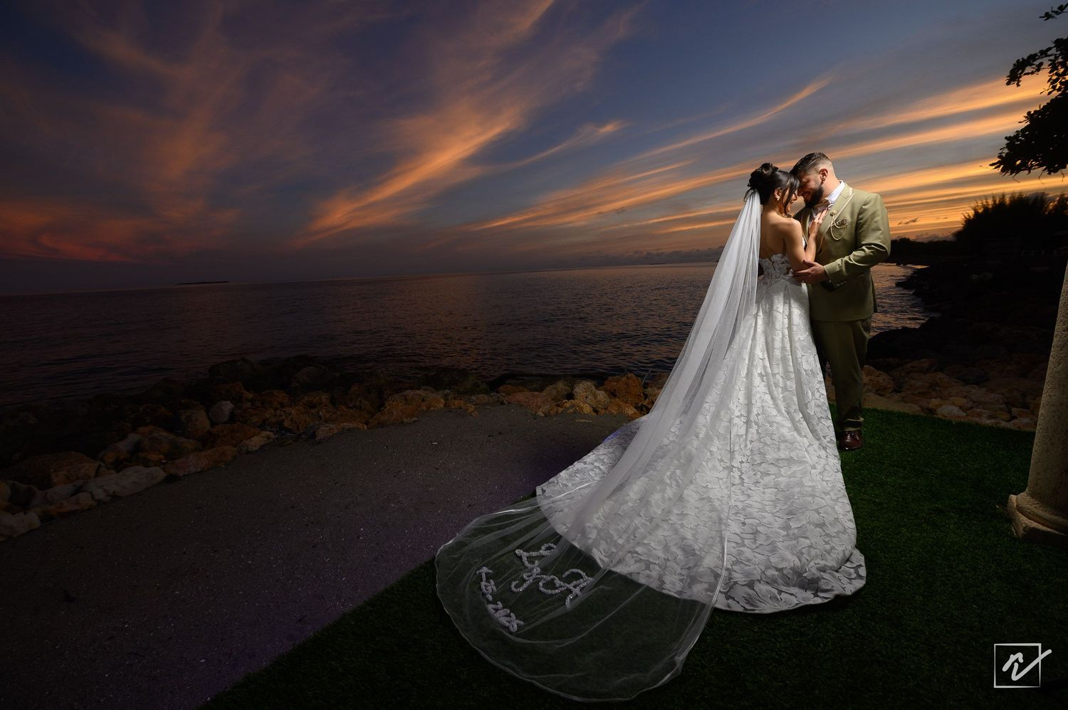 Bride in a long lace gown and veil embraces her groom on a grassy shoreline beneath a vivid orange and purple sunset sky over the water.