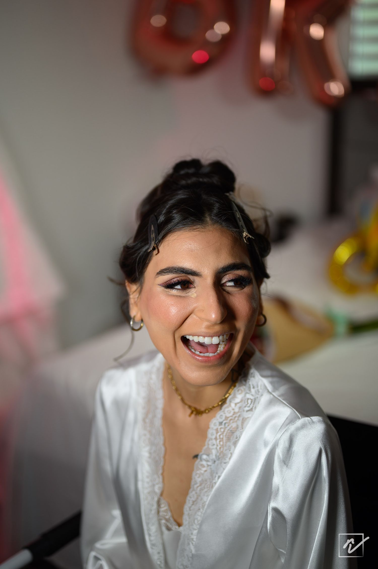 Smiling bride in a white satin robe with lace trim, styled updo and glowing makeup, laughing while getting ready on her wedding day.