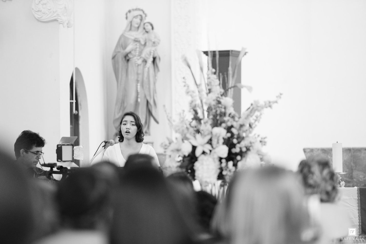 Black and white photo of a woman speaking at a church podium with a Virgin Mary statue and floral arrangements behind her.