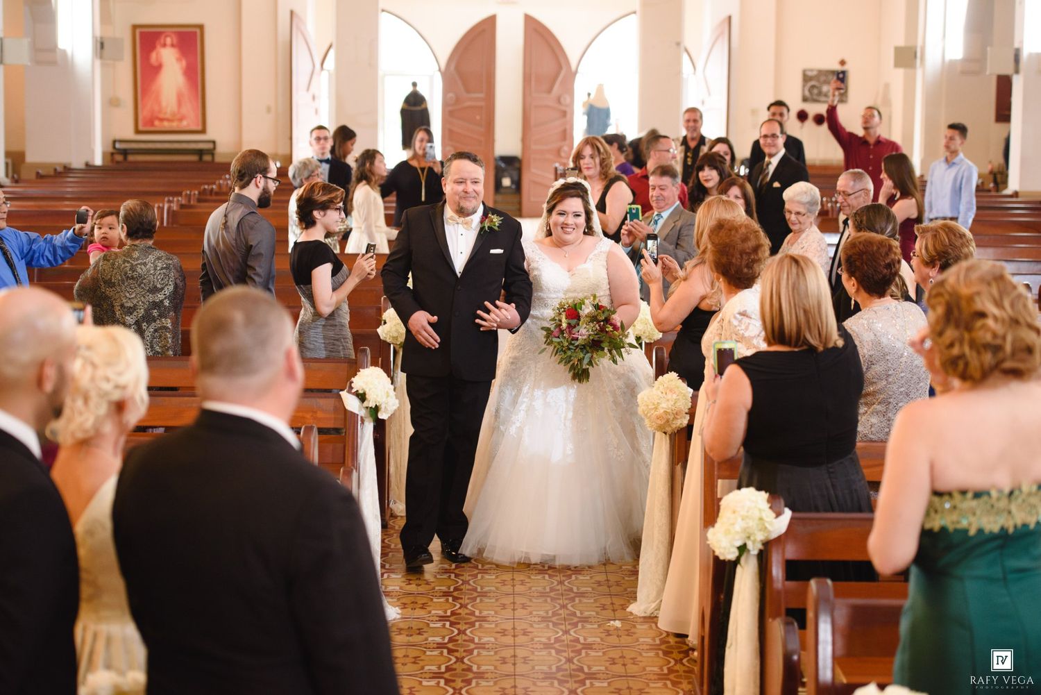 Bride and groom walk down the aisle of a decorated church surrounded by smiling wedding guests.