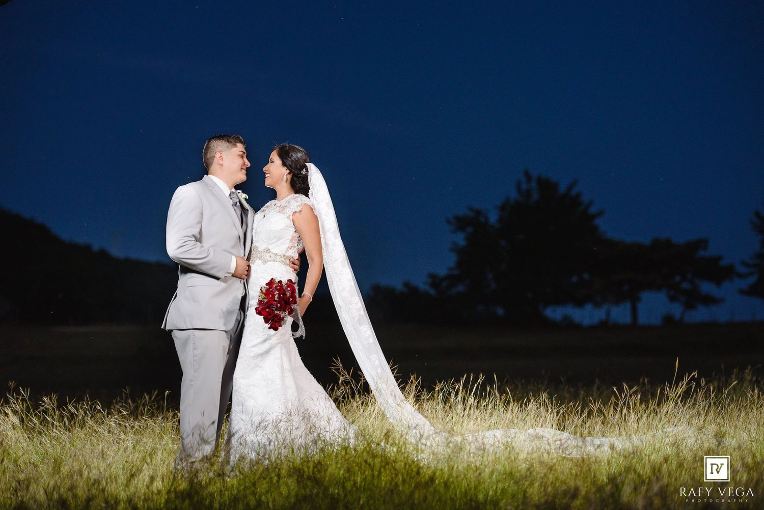 Bride and groom share a romantic moment in a moonlit field at night, with the bride holding a red bouquet.