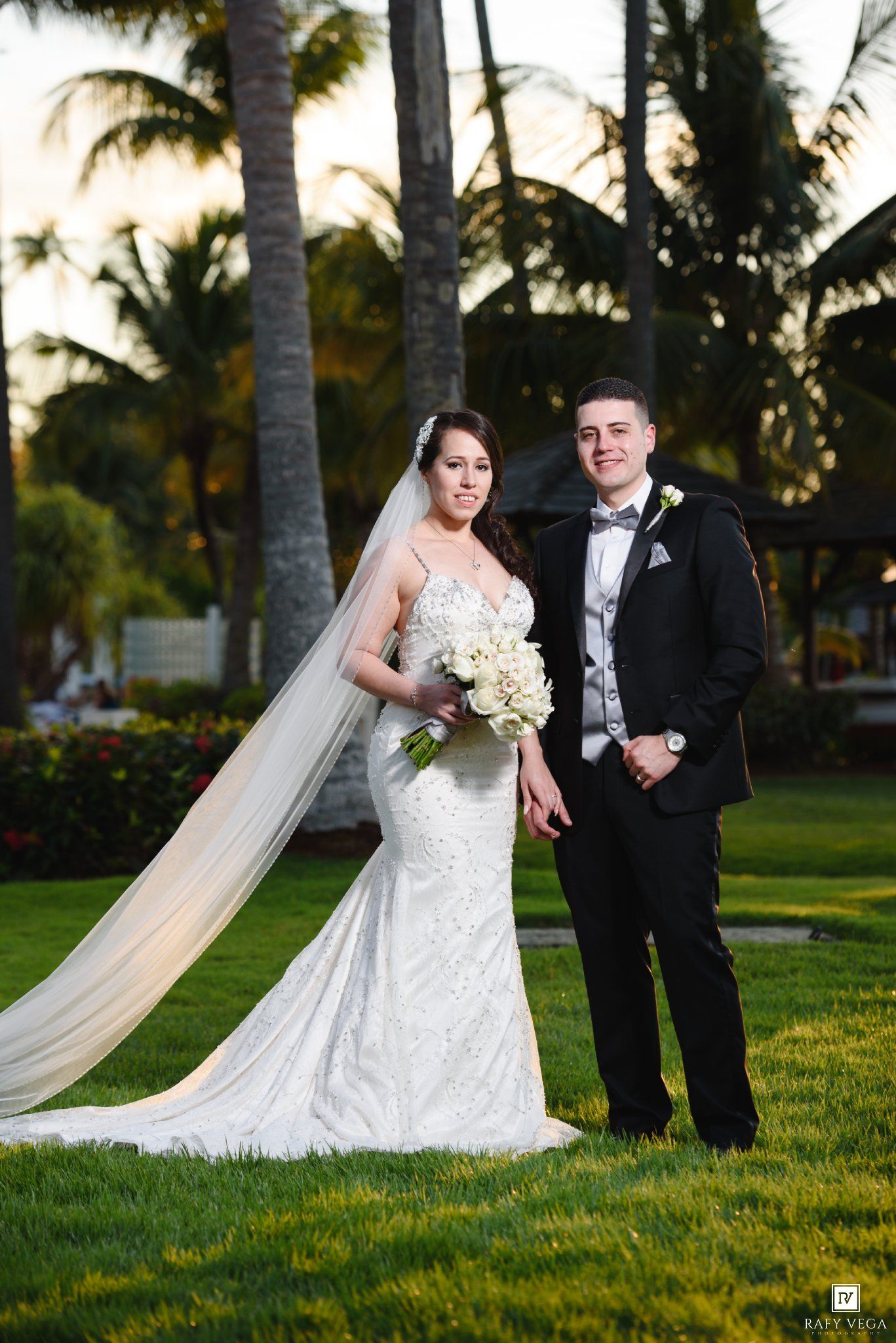 Bride in white mermaid gown with veil and groom in black tuxedo pose on green lawn with palm trees at sunset.