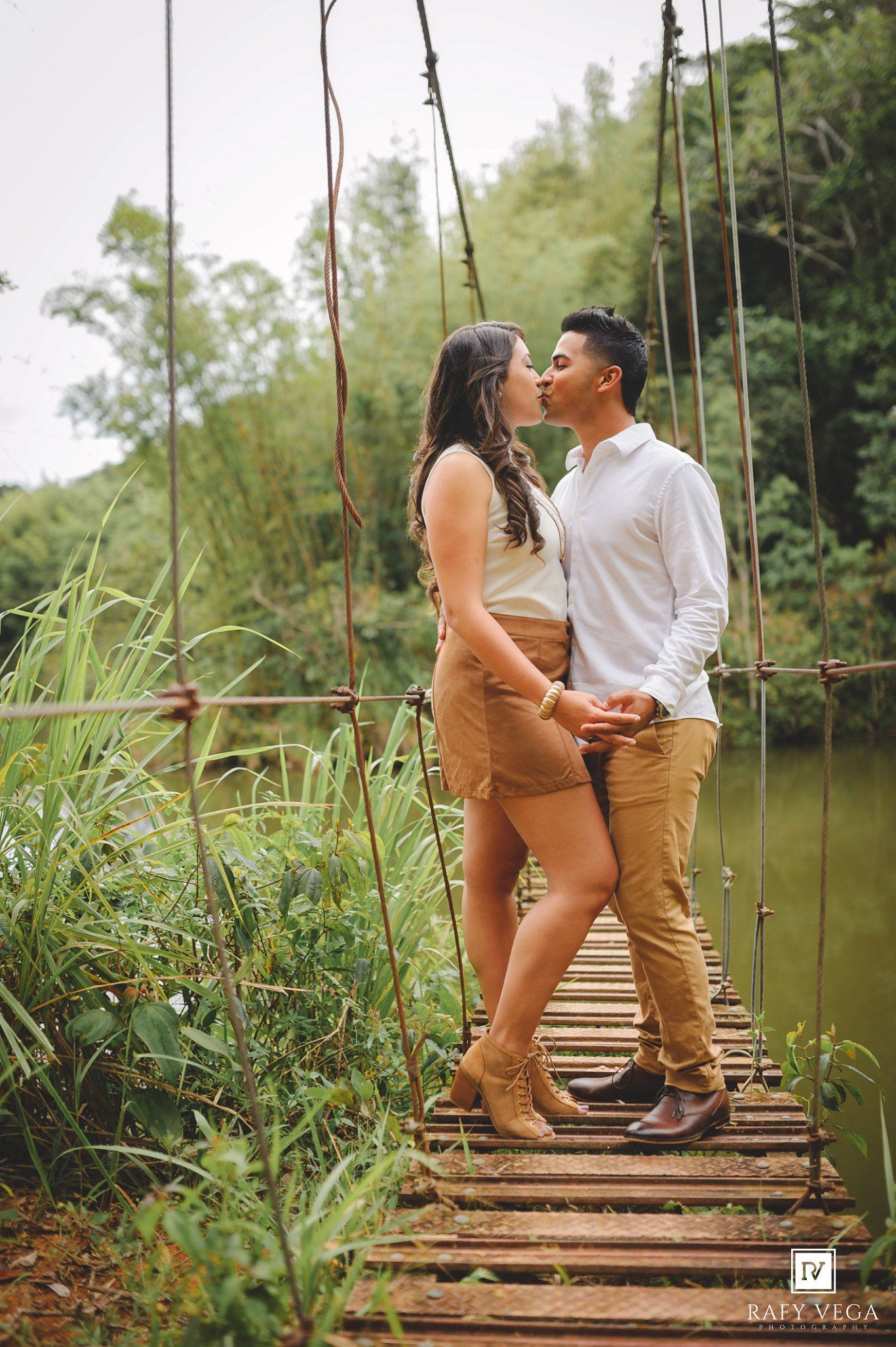 Couple sharing a romantic moment on a rustic wooden suspension bridge surrounded by lush green trees and a river.
