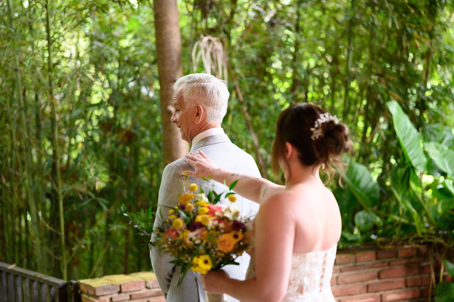 Bride holding colorful bouquet touches groom's shoulder in lush garden setting during outdoor wedding ceremony.