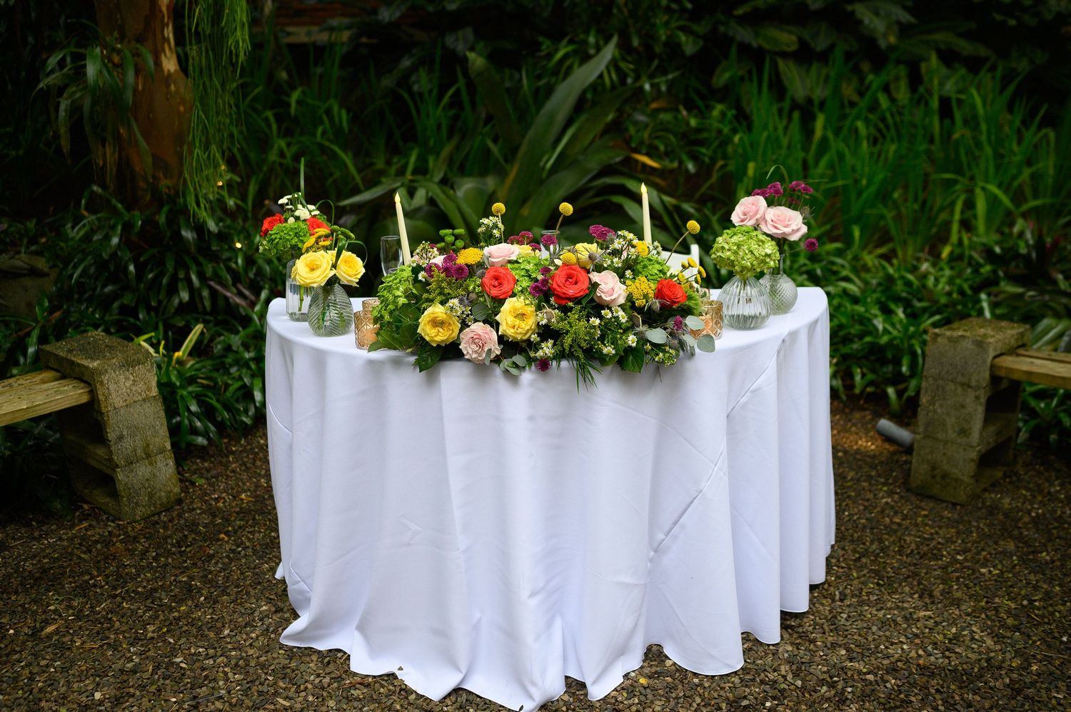 Elegant wedding sweetheart table with white linen and colorful floral centerpiece in lush garden setting.
