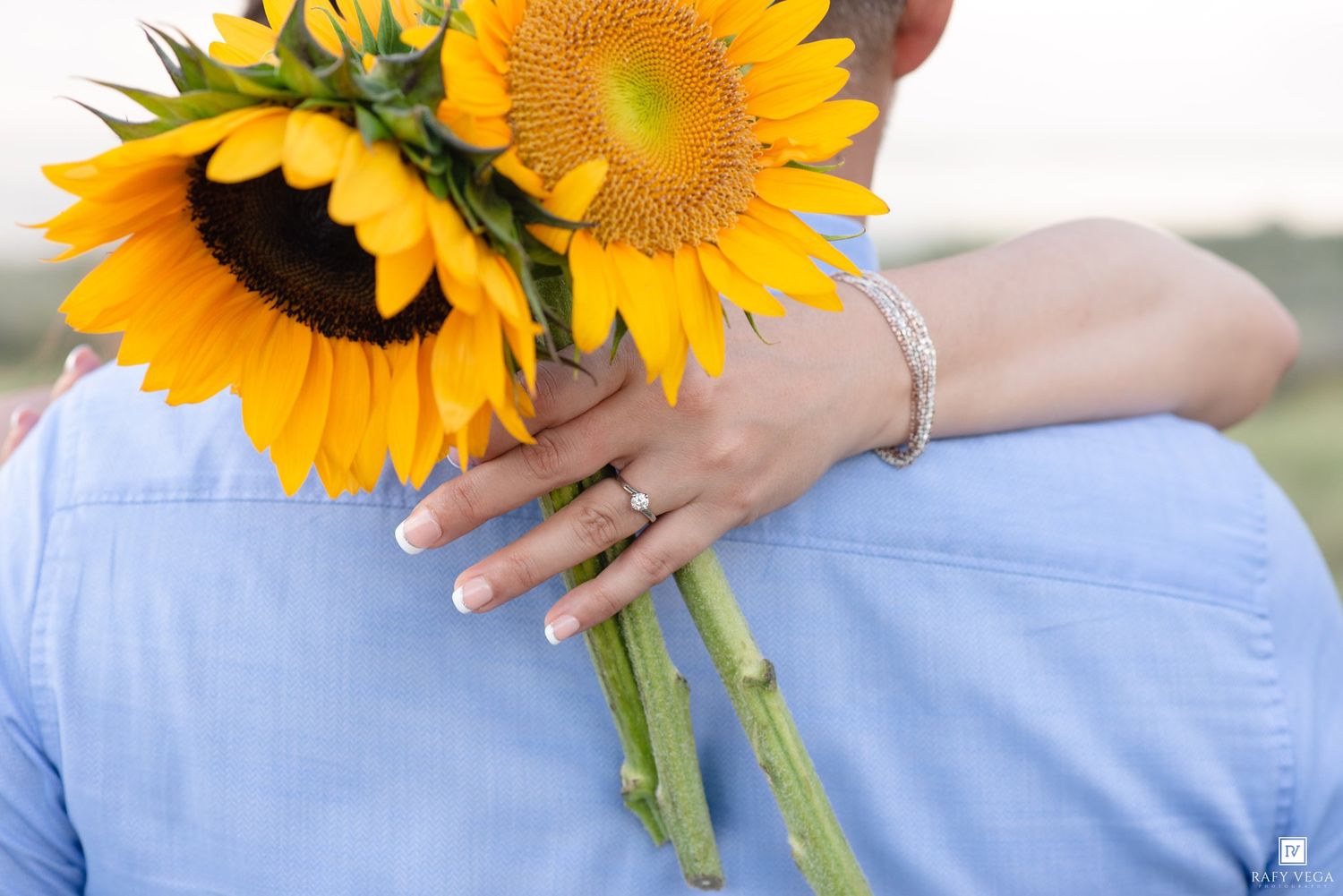 Couple embracing outdoors, woman holding bright yellow sunflowers, wearing bracelet and ring, man in light blue shirt.