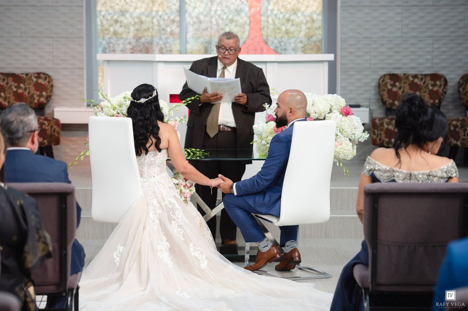 Bride and groom hold hands during indoor wedding ceremony while officiant reads vows at elegant white altar.