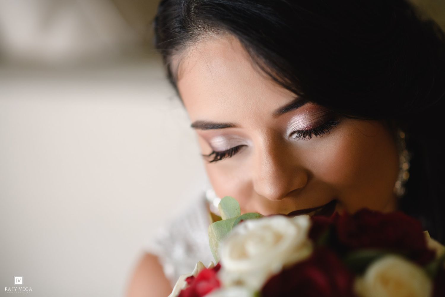 Bride smelling a bouquet of red and white roses, wearing pearl earrings with elegant bridal makeup.