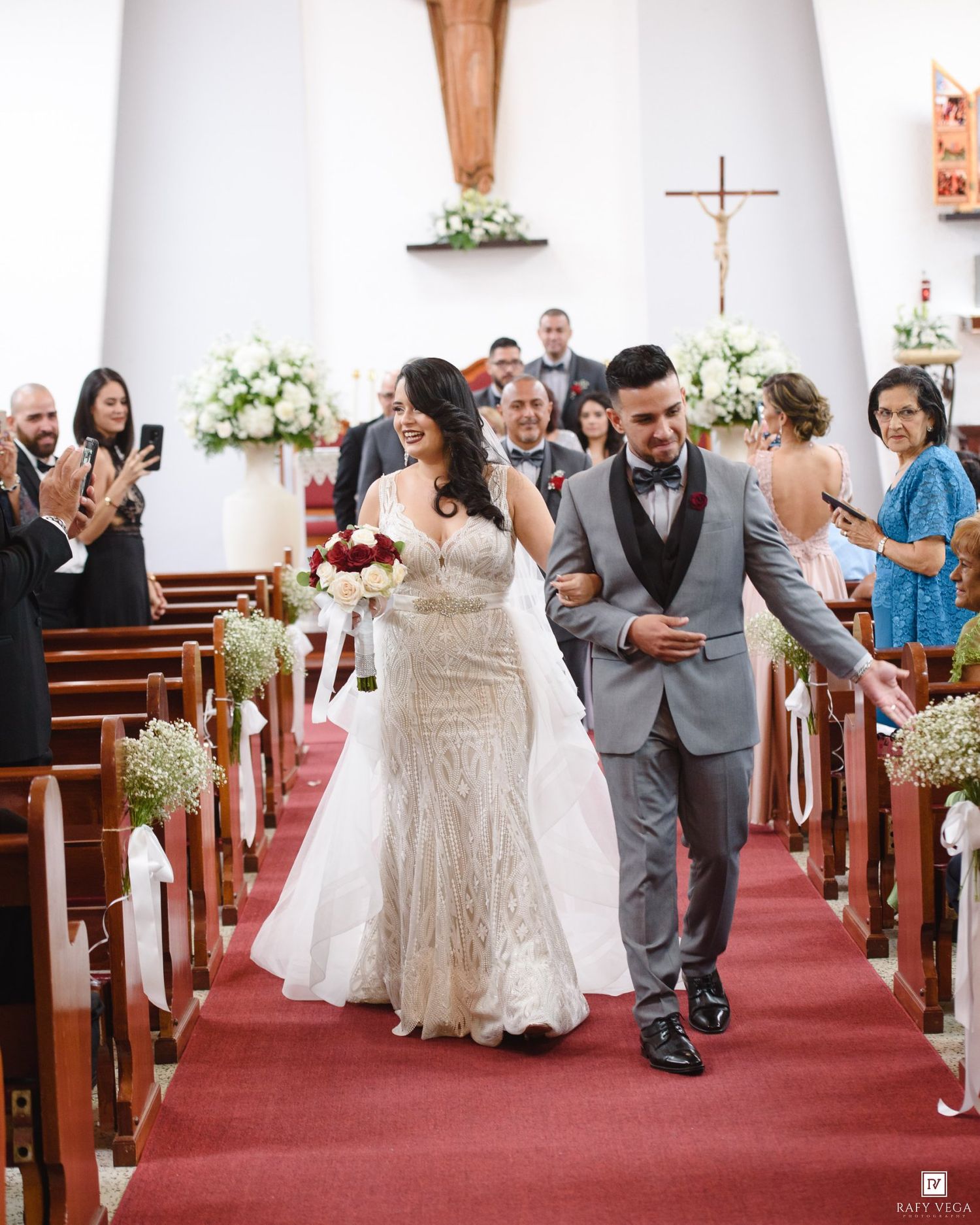 Bride and groom walk down church aisle after wedding ceremony, bride holding red bouquet, groom in gray suit.