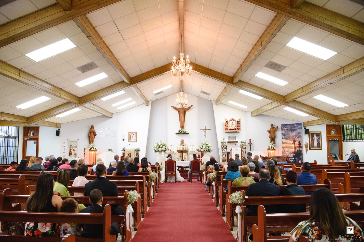 Church wedding ceremony with guests seated in wooden pews along red carpet aisle toward floral altar.