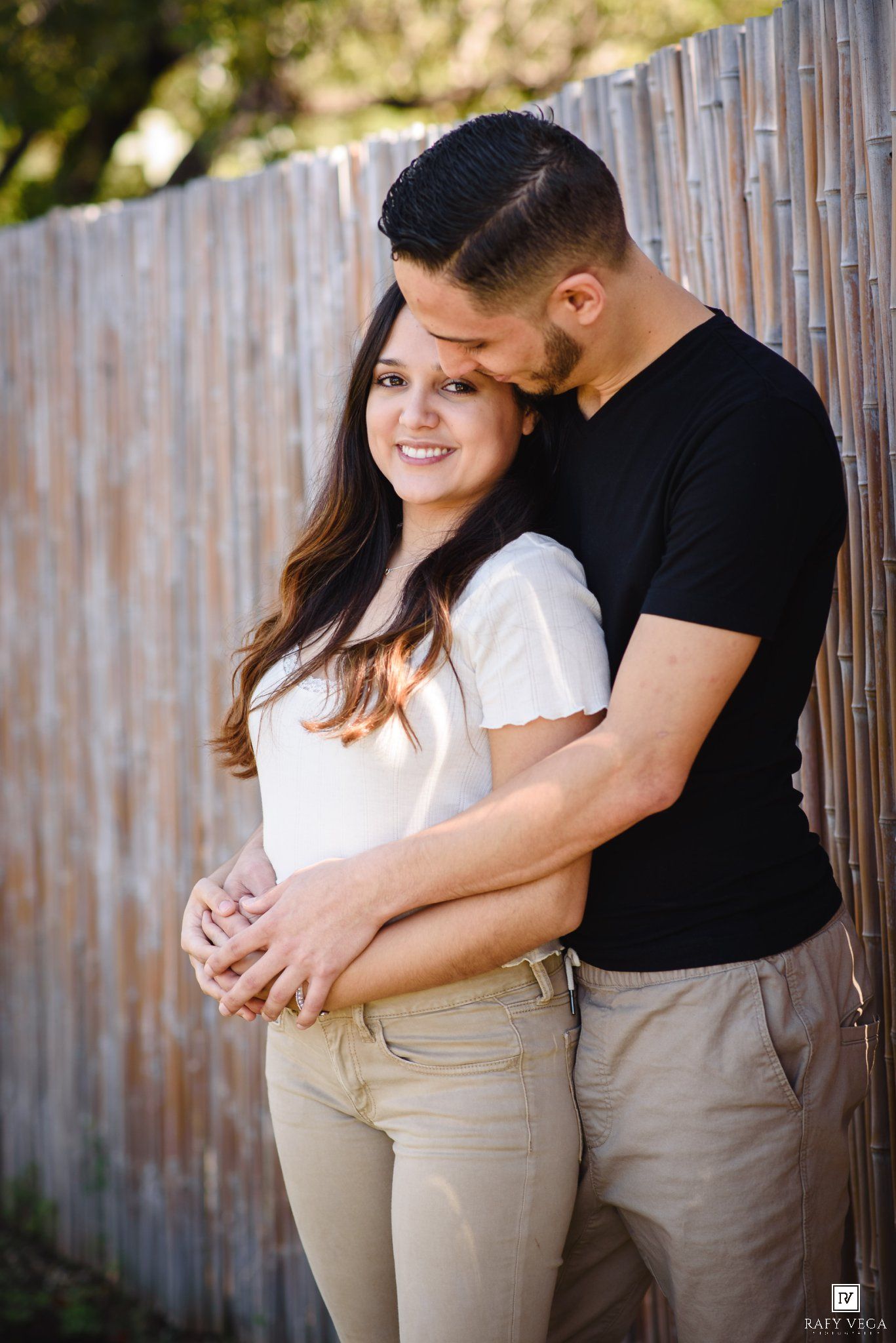 A couple embraces lovingly against a bamboo fence, the man kissing her head while she smiles warmly in a white top.