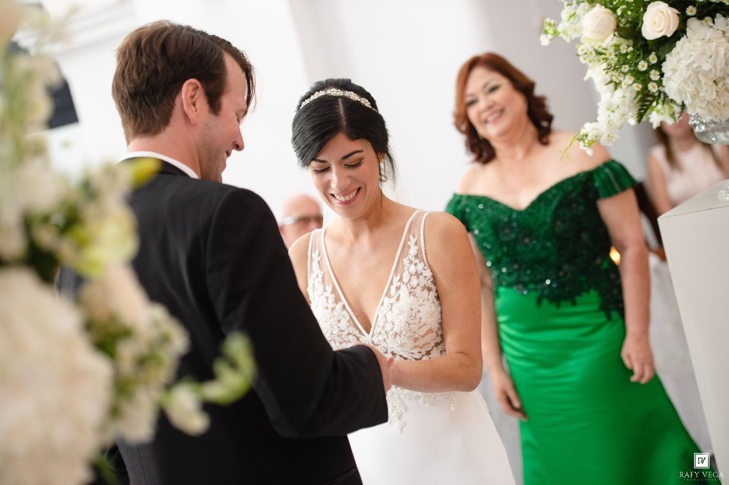 Bride in lace gown holds grooms hands as woman in green dress looks on.