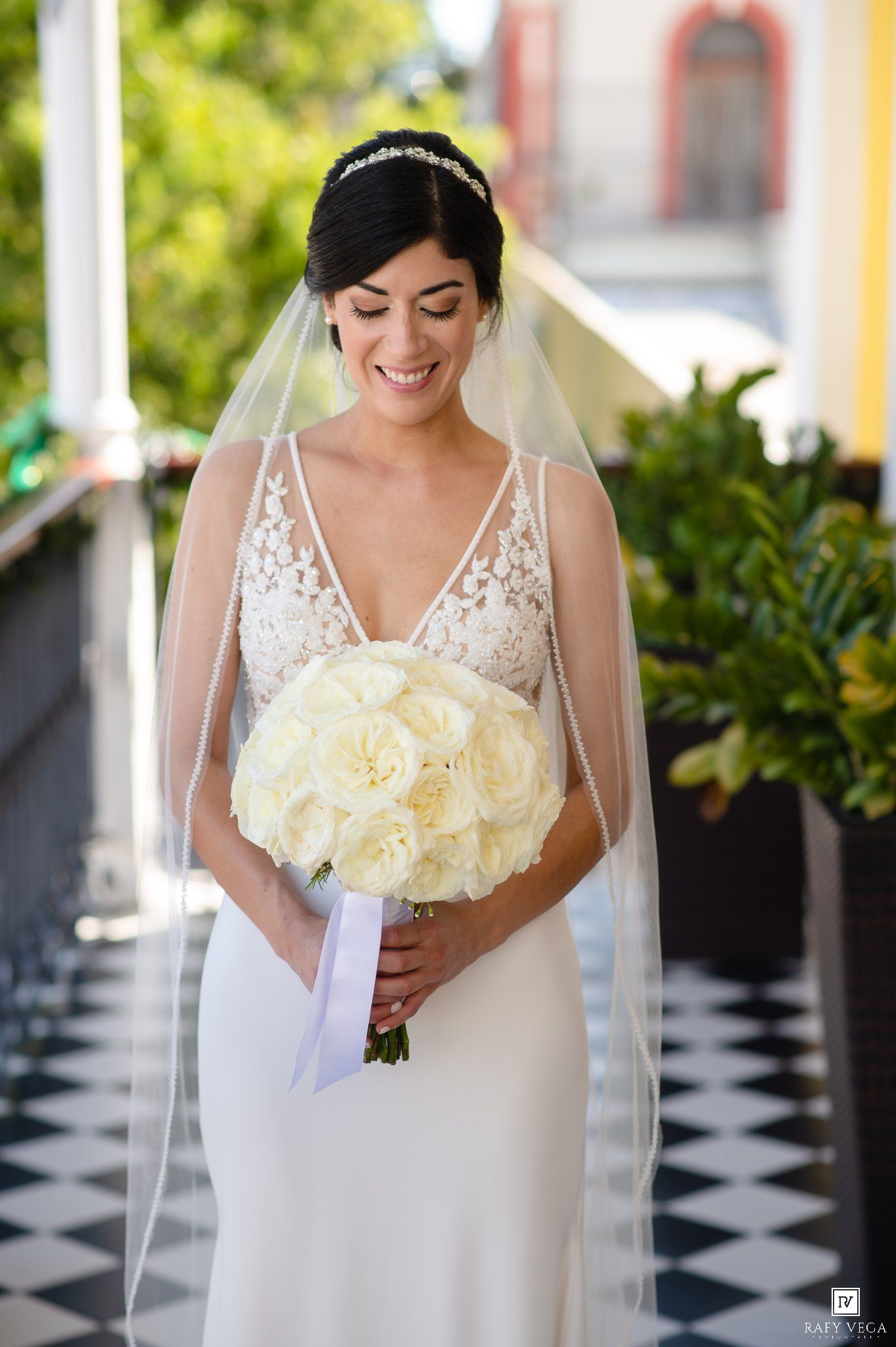 Smiling bride in lace wedding dress holds white rose bouquet, wearing veil and tiara on checkered floor.