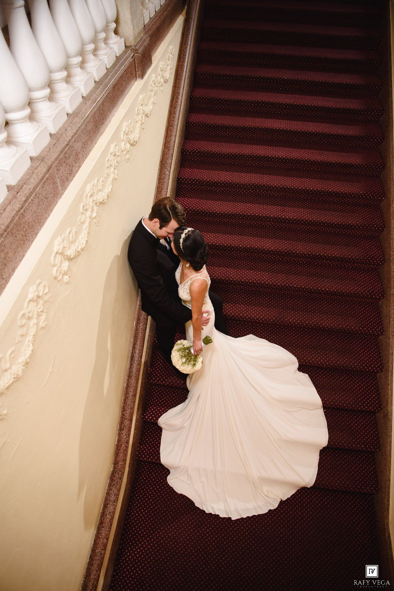 Aerial view of bride and groom sharing romantic moment on grand staircase with red carpet and ornate white railing.
