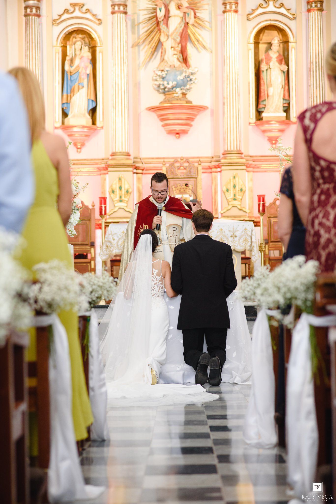 Bride and groom kneeling at church altar during Catholic wedding ceremony with ornate golden decor.