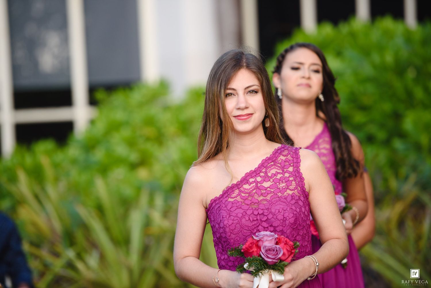 Bridesmaid in purple lace one-shoulder dress holding bouquet at outdoor wedding ceremony.
