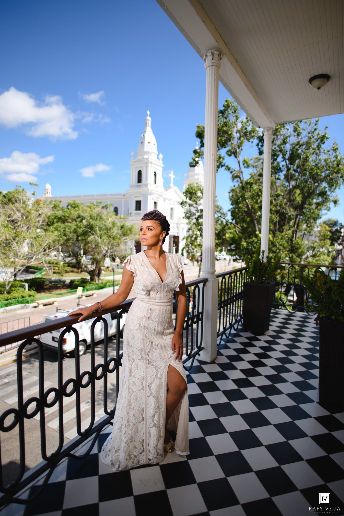 Woman in white lace gown poses on checkered balcony with white church and blue sky backdrop.