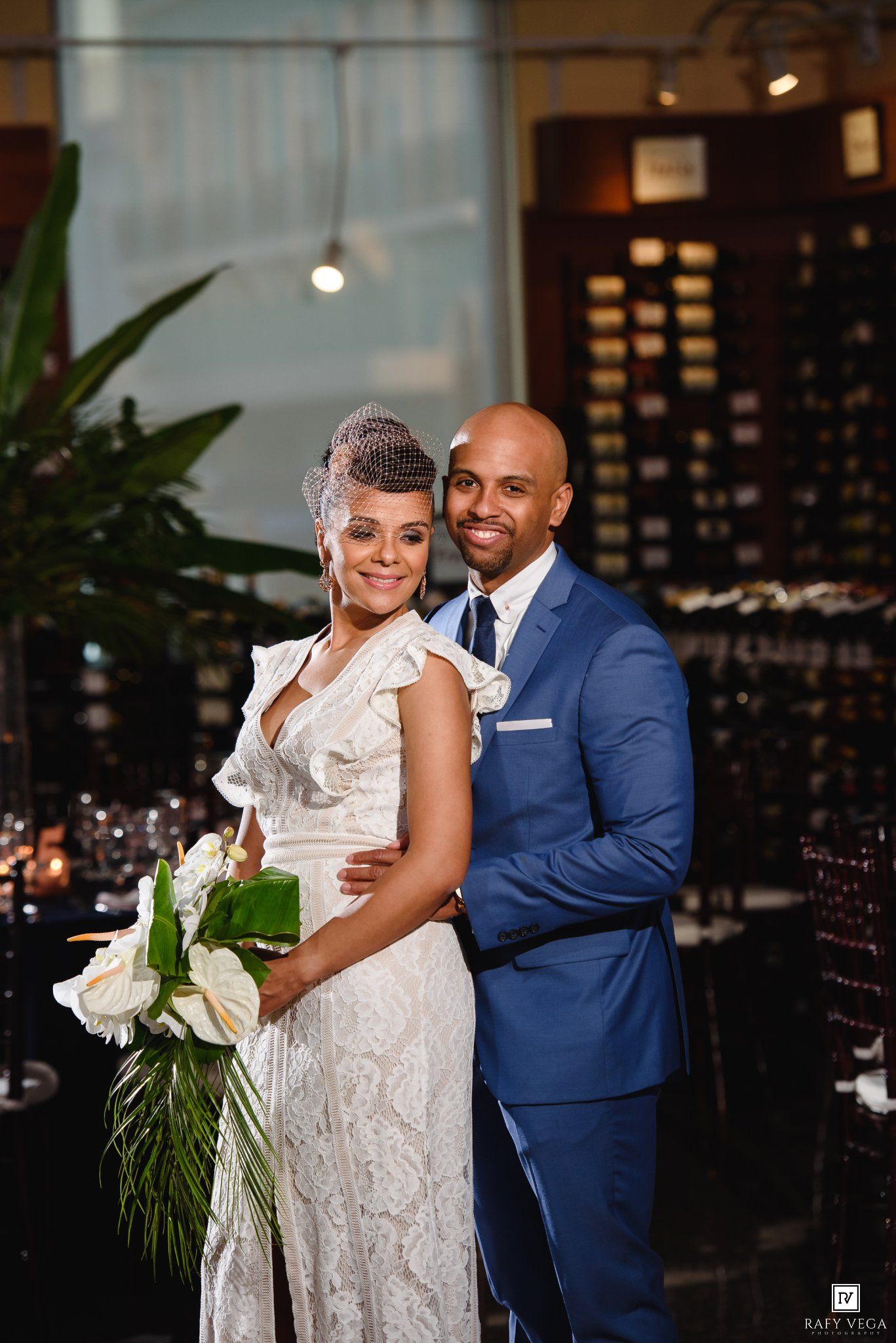 Elegant couple posing at wedding reception, bride in white lace gown holding bouquet, groom in blue suit, wine cellar backdrop.