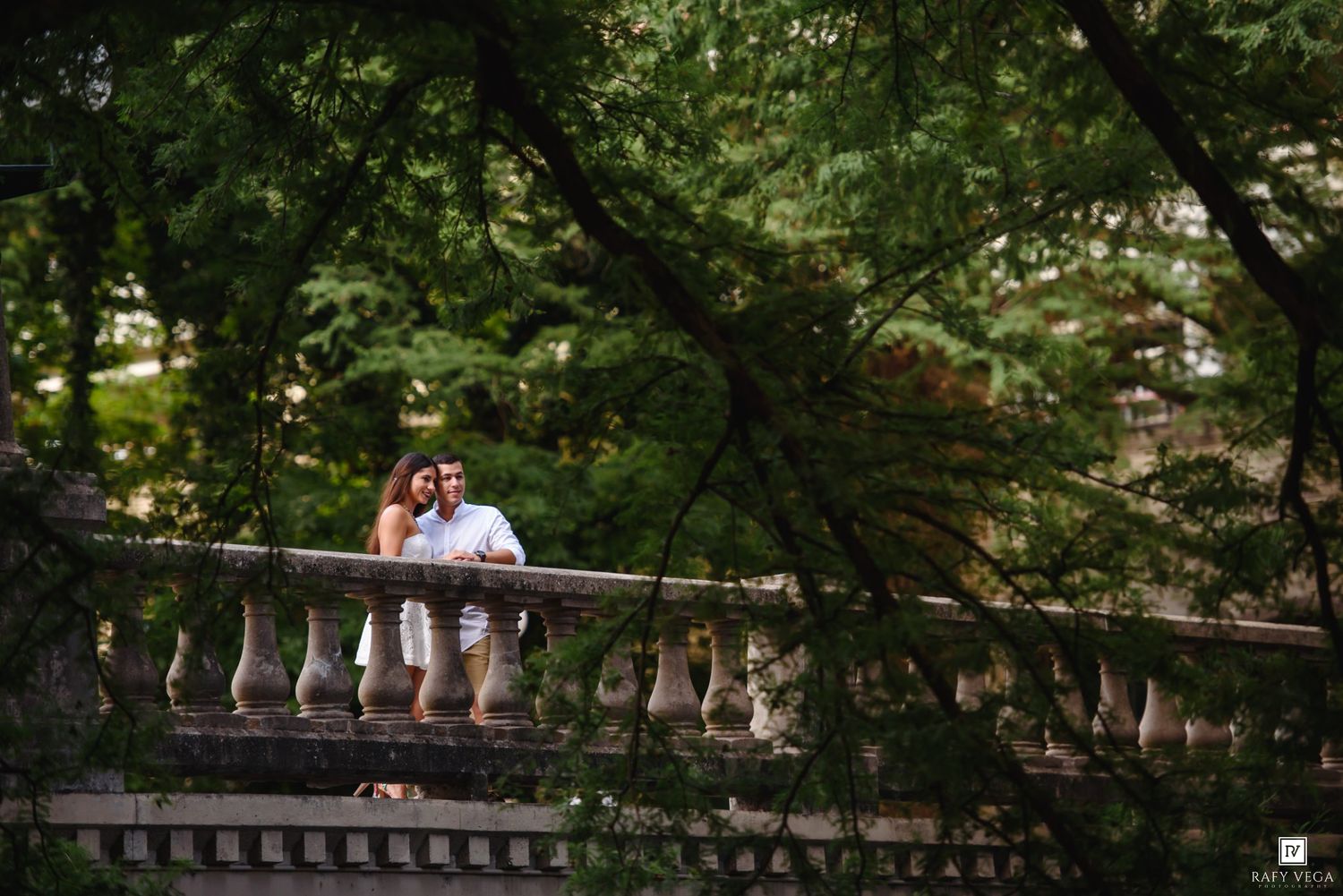 Couple embracing on a stone balustrade bridge surrounded by lush green trees in a romantic outdoor setting.