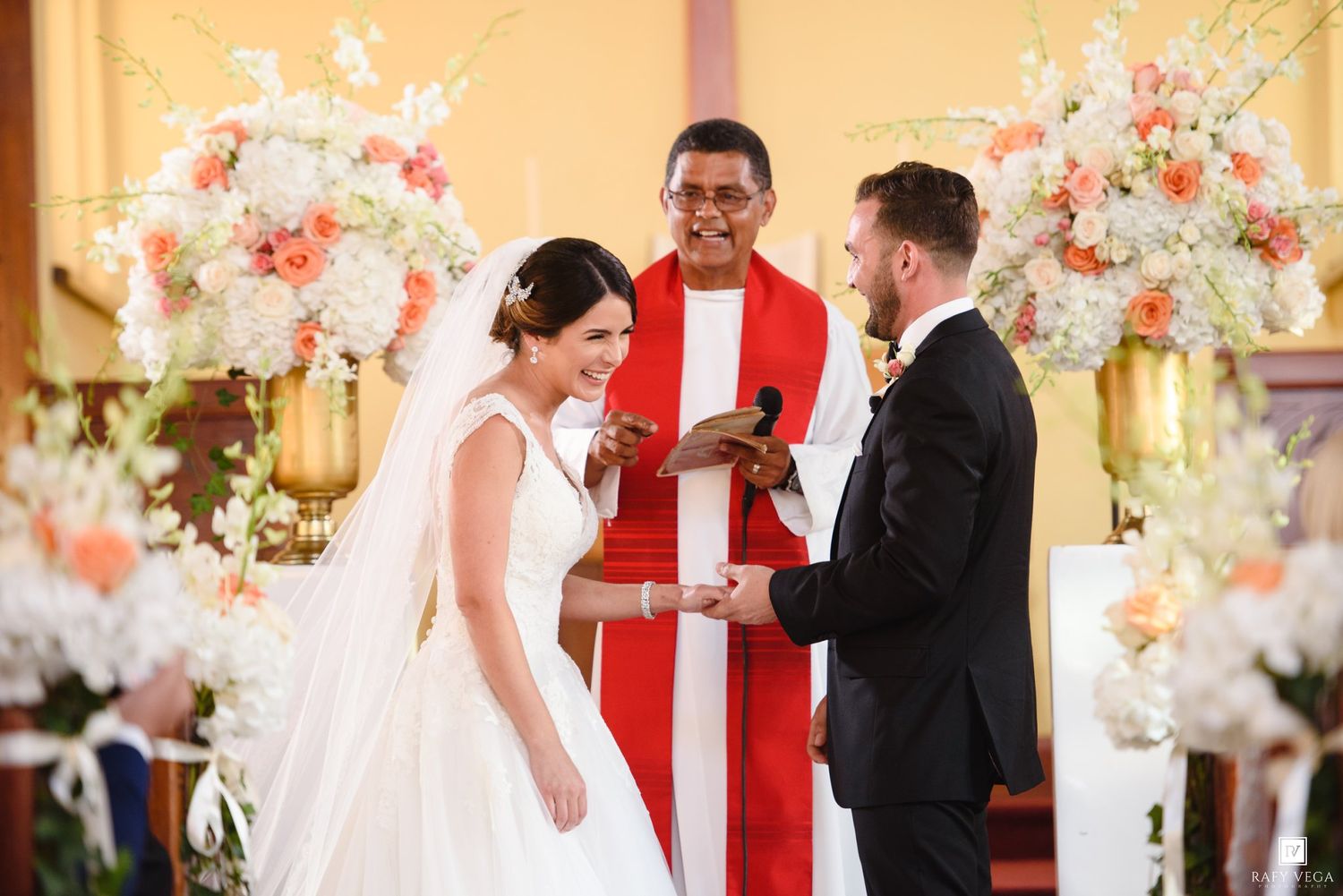 Bride and groom exchange vows at church altar with priest in red vestments and floral arrangements.