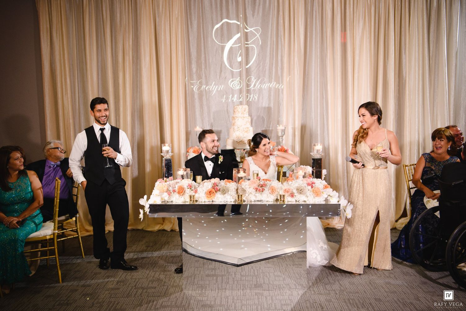 Wedding party toasting newlyweds at decorated sweetheart table during elegant indoor reception ceremony.