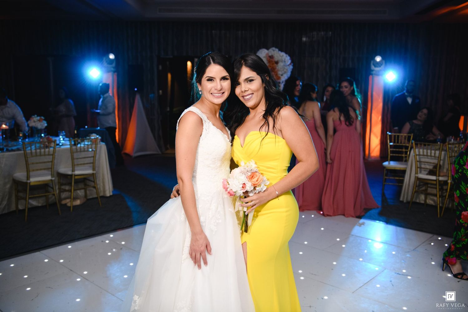 A bride in white gown poses with bridesmaid in yellow dress on a white dance floor at wedding reception.