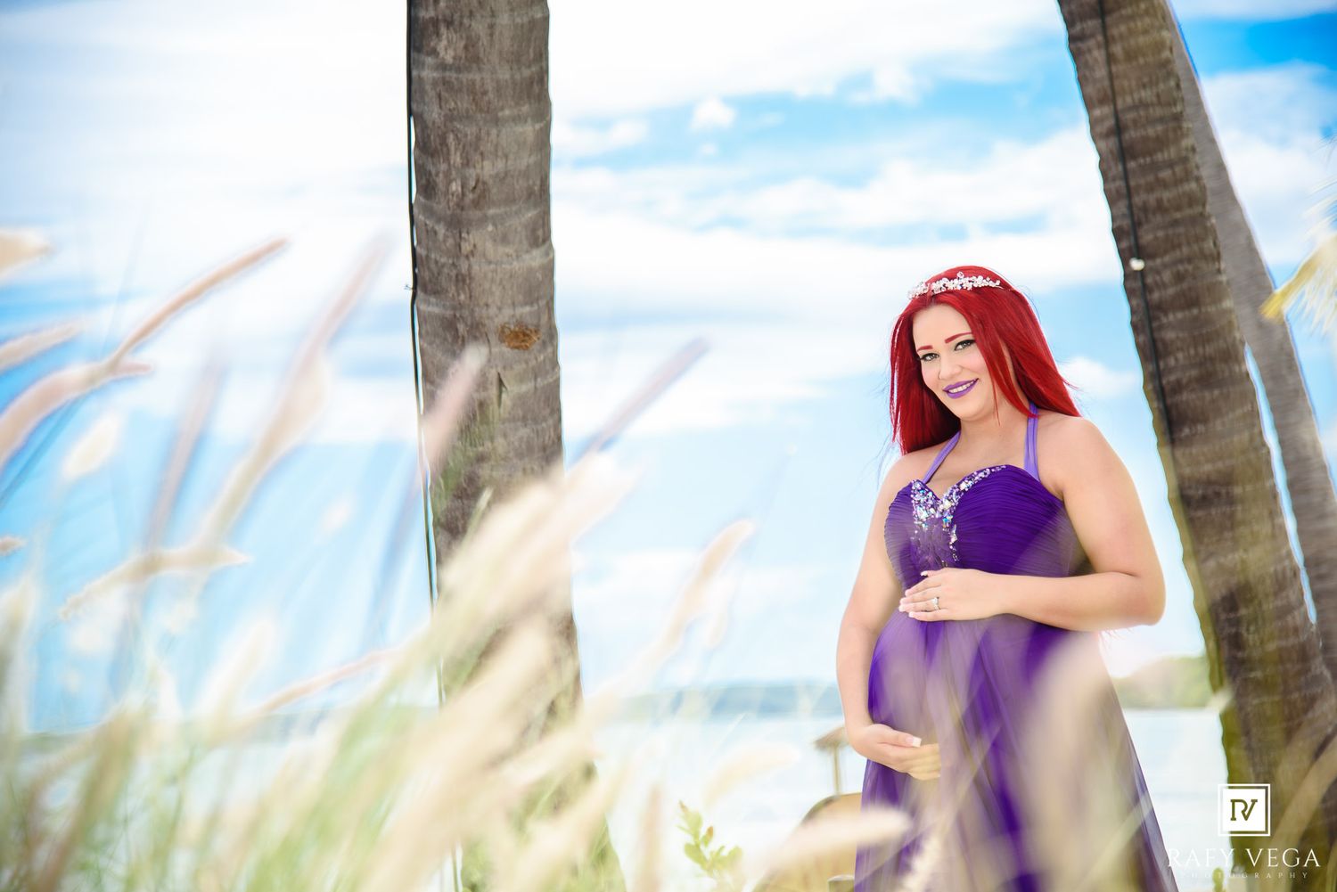 Pregnant woman with red hair in purple dress posing near palm trees on a sunny beach.